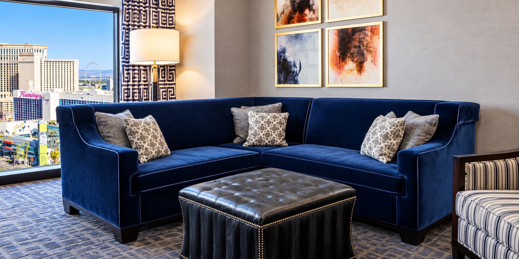 A blue sectional sofa with patterned cushions, a leather ottoman, and a view of a cityscape through a large window.