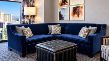A blue sectional sofa with patterned cushions, a leather ottoman, and a view of a cityscape through a large window.