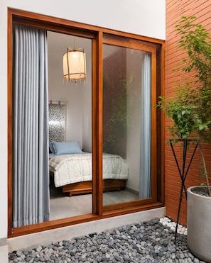 Quarto com porta de vidro deslizante, cama com colcha estampada, luminária pendente e planta em vaso.
