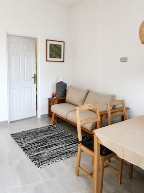 Quarto com porta branca, sofá bege, cadeira de madeira, mesa pequena, tapete preto e branco e quadro na parede.