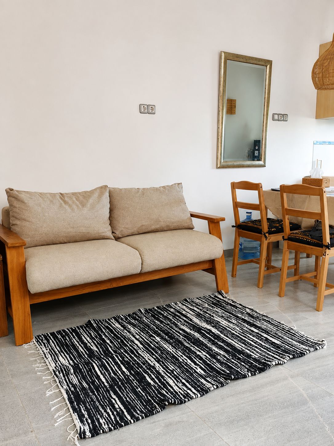 A minimalist living room with a sofa, a dining table with chairs, a black and white rug, and a mirror on the wall.