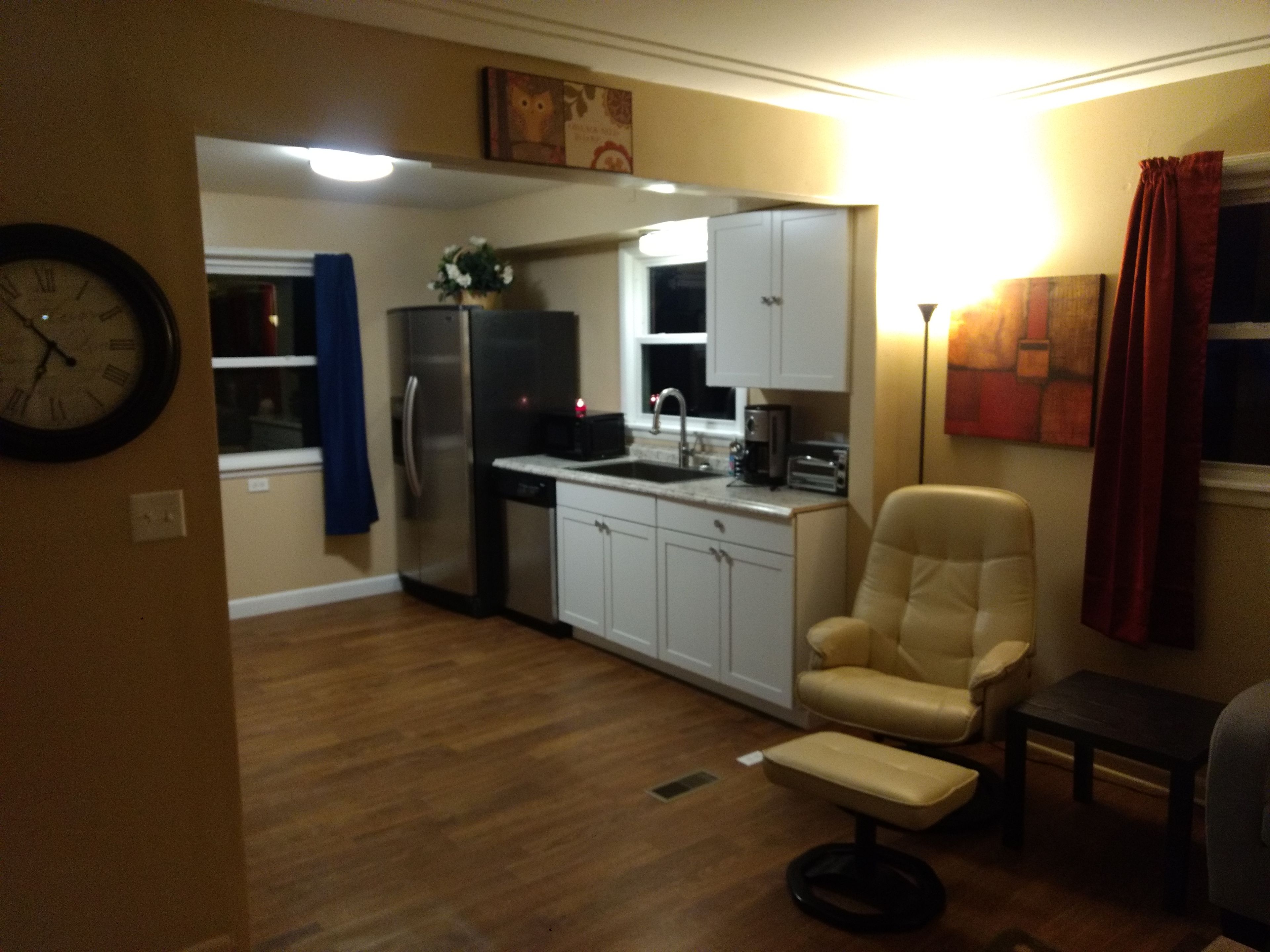 A compact kitchen with white cabinets, a refrigerator, and a sink. There is a beige armchair and a small table in the living area. A clock is mounted on the wall.