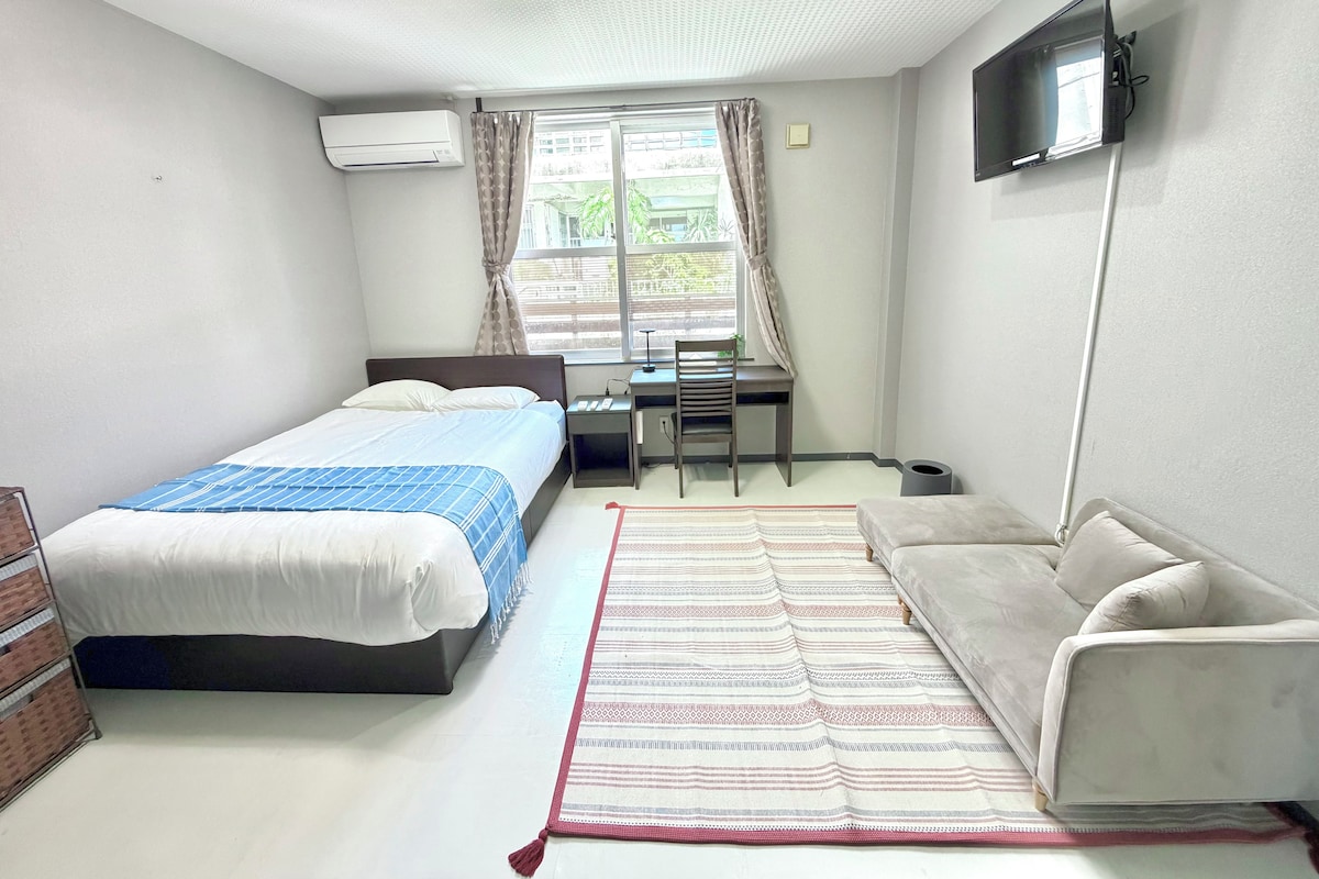 Un dormitorio con cama, escritorio, silla, sofá, televisor y ventana con cortinas.