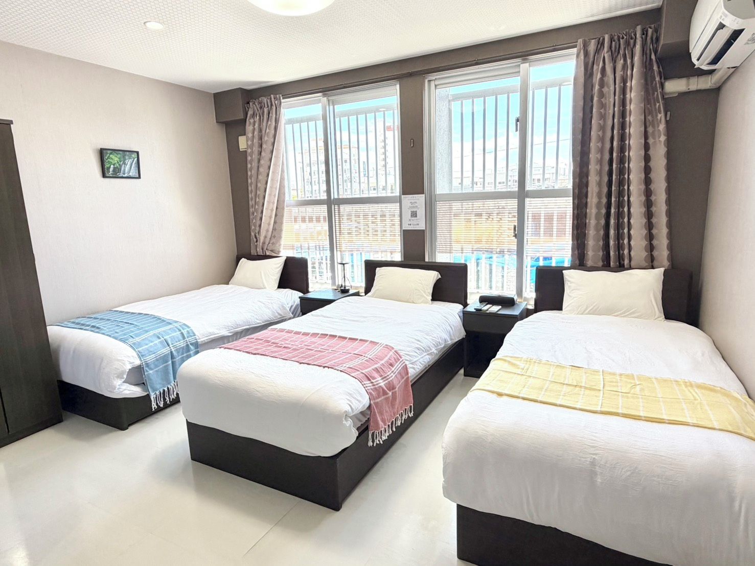 A hotel room with three beds, large windows with curtains, an air conditioning unit, and a small picture on the wall.