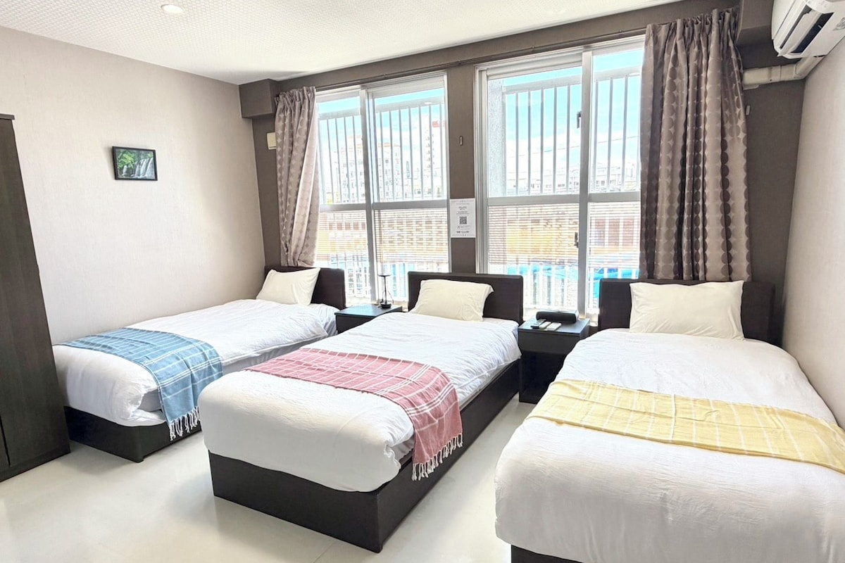 Habitación de hotel con tres camas, ventanas grandes con cortinas, un equipo de aire acondicionado y un pequeño cuadro en la pared.