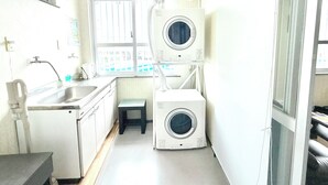 Een kleine wasruimte met een wasbak, twee gestapelde wasmachines en een raam met jaloezieën.
