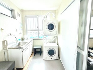 A compact laundry room with a sink, two stacked washing machines, and a window with blinds. - ishigaki bus terminal inn (Ishigaki)