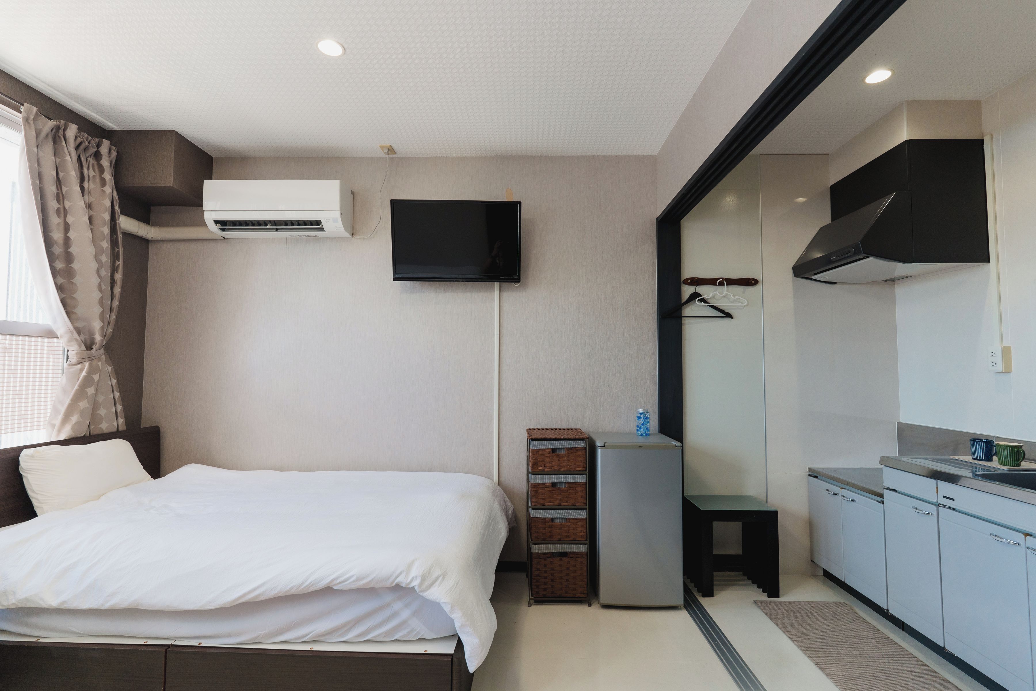 A hotel room with a bed, an air conditioning unit, a television, a small refrigerator, a mirror, and a kitchenette.