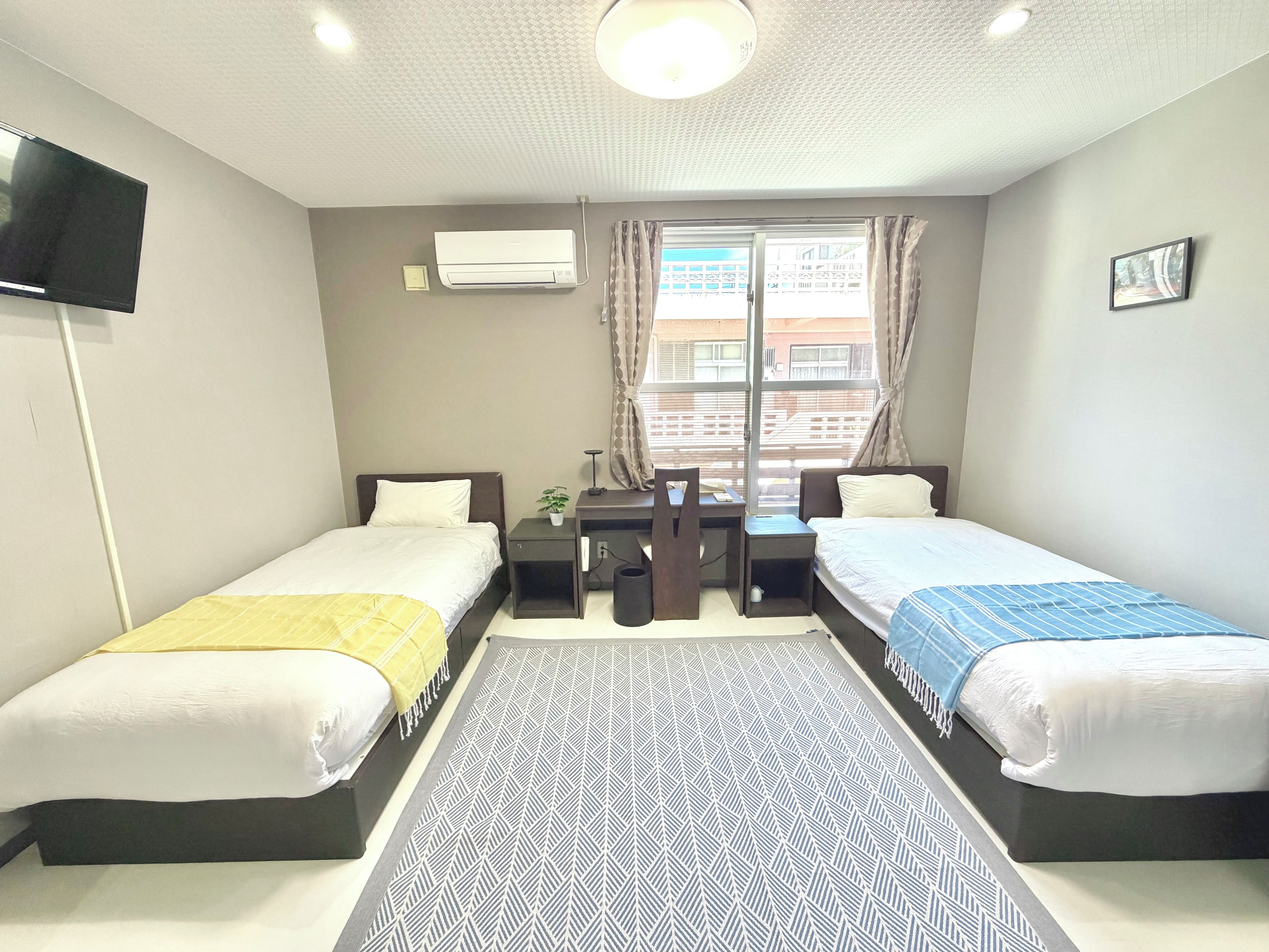 A hotel room with two beds, a desk, a chair, a TV, an air conditioning unit, and a window with curtains.