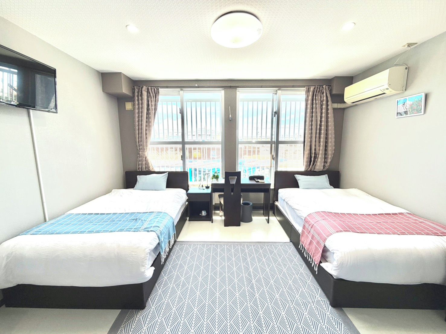 A hotel room with two beds, a desk, a chair, and a view of the city through large windows.