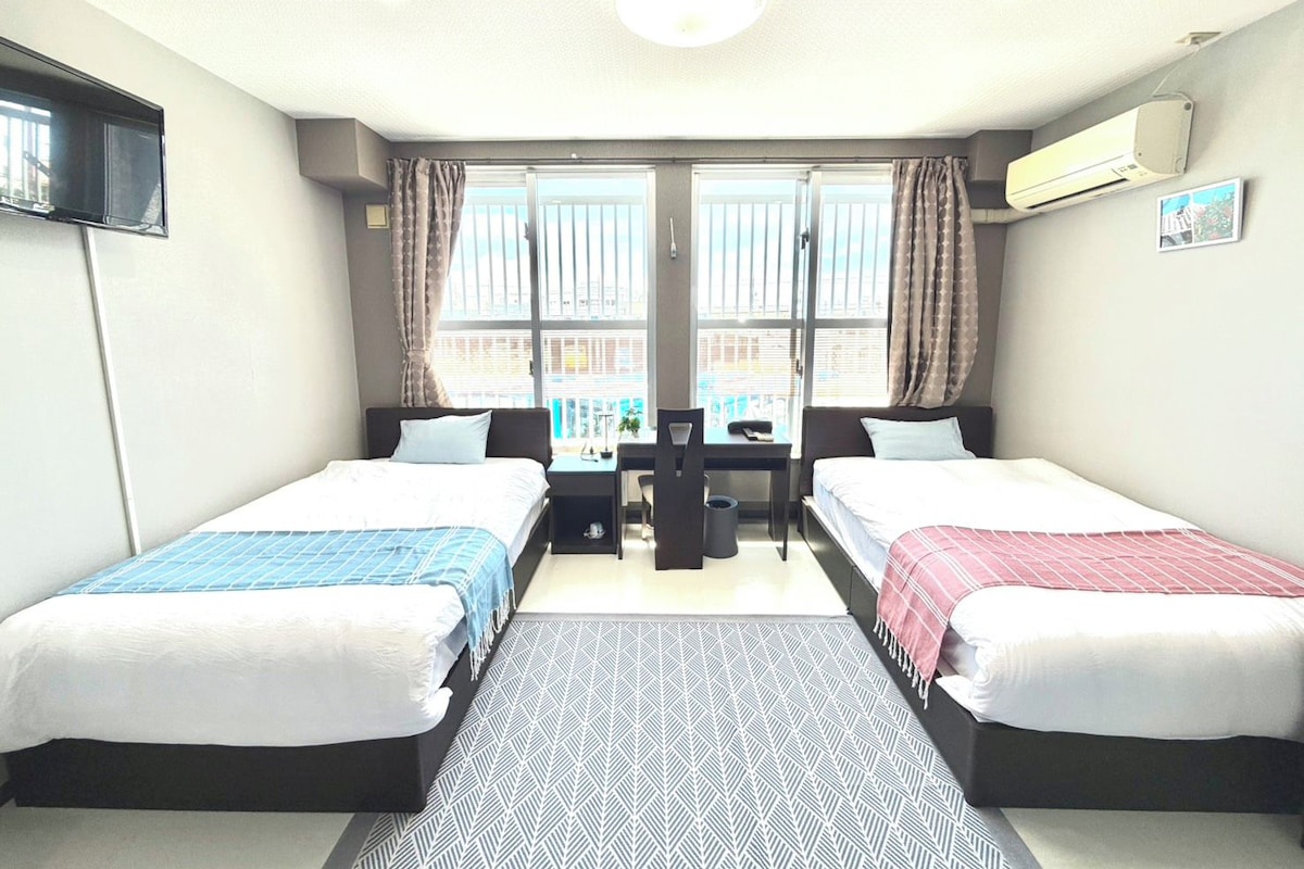 Habitación de hotel con dos camas, un escritorio, una silla y vistas de la ciudad a través de amplios ventanales.
