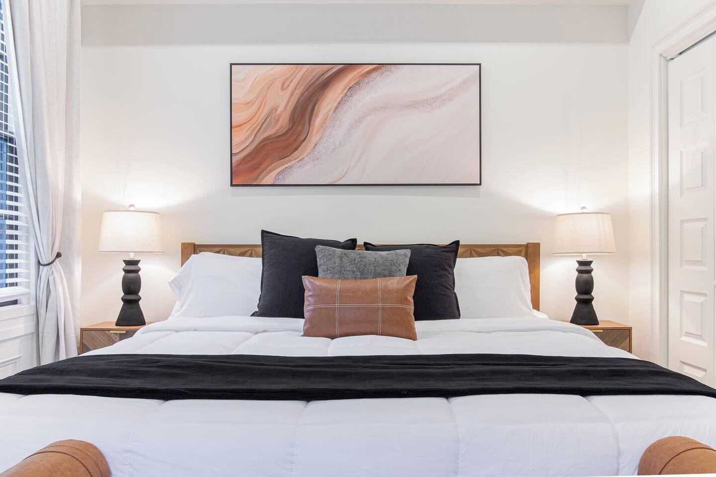 A neatly made bed with a black bedspread and multiple pillows, a marble-like artwork above it, and two bedside lamps on either side.