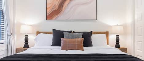 A neatly made bed with a black bedspread and multiple pillows, a marble-like artwork above it, and two bedside lamps on either side.