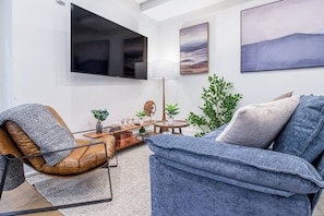Ein modernes Wohnzimmer mit einem Ledersessel, einem blauen Sofa, einem Flachbildfernseher, einer Lampe und einem Gemälde an der Wand.