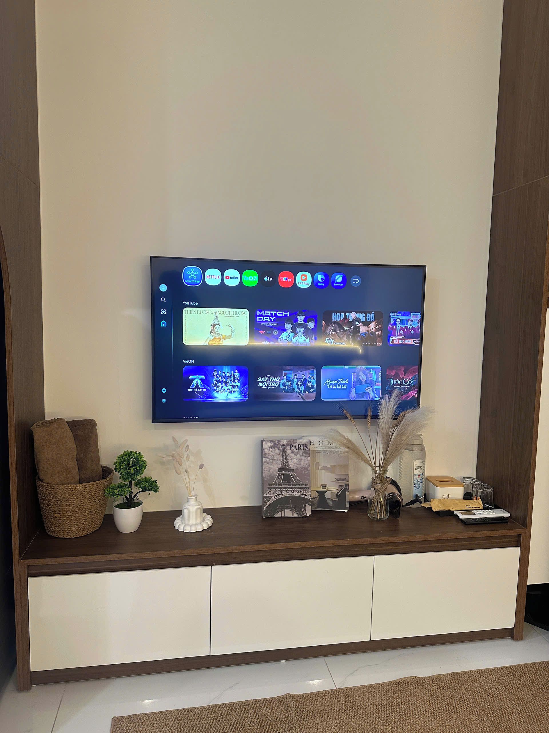 Un televisor de pantalla plana montado en una pared con una pantalla dividida, un mueble blanco debajo y un estante decorativo con una planta, un jarrón y un libro.