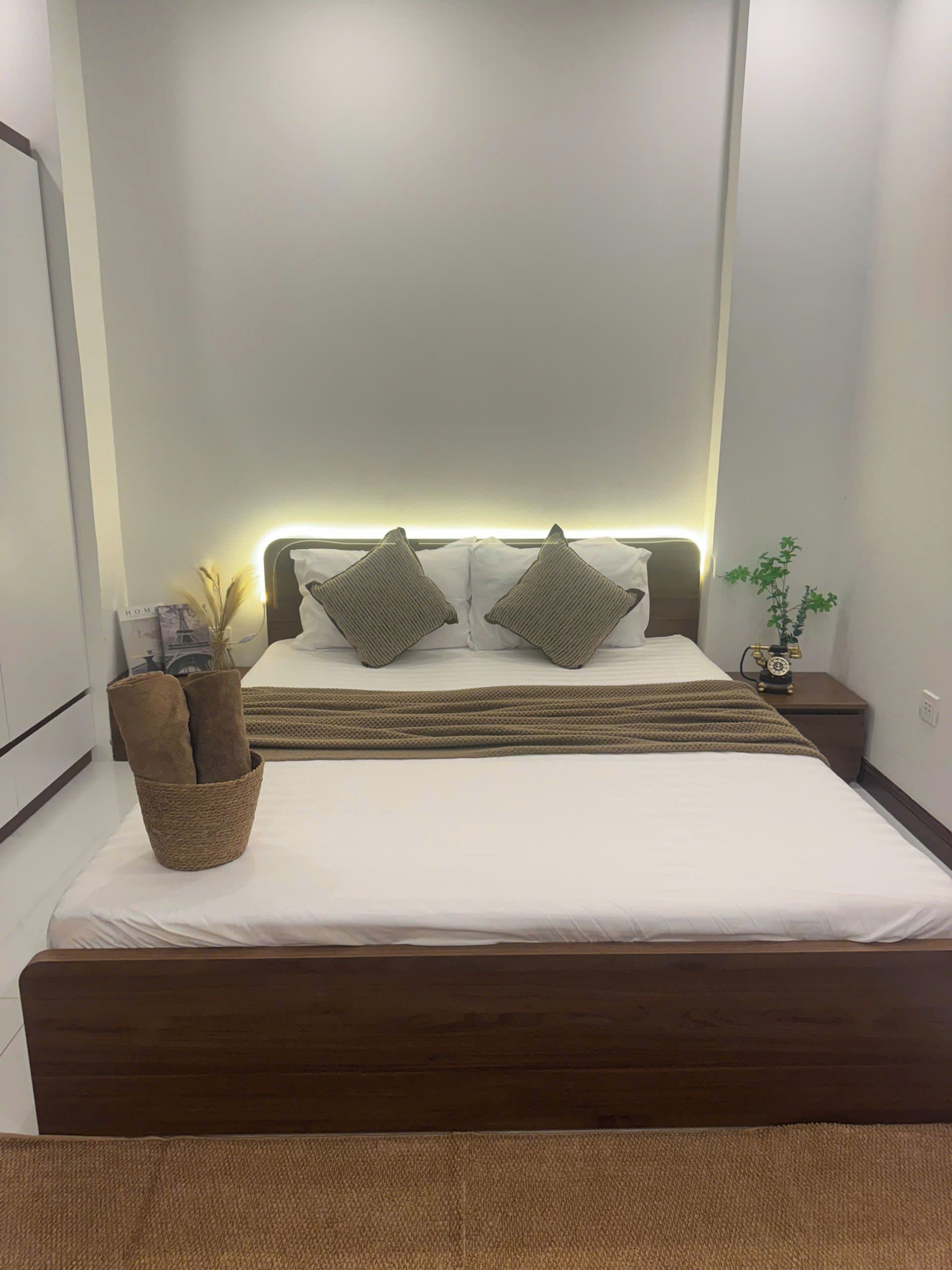 A neatly made bed with a headboard, two pillows, a bedside table with a clock, and a small plant.
