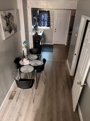 Ein Essbereich mit Tisch, Stühlen und einer Vase mit einer Blume. Im Hintergrund ist eine Küche mit einem Fenster und Vorhängen zu sehen.