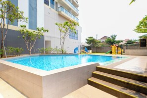 A swimming pool with steps leading into it, surrounded by a building with multiple floors and a playground area with slides.