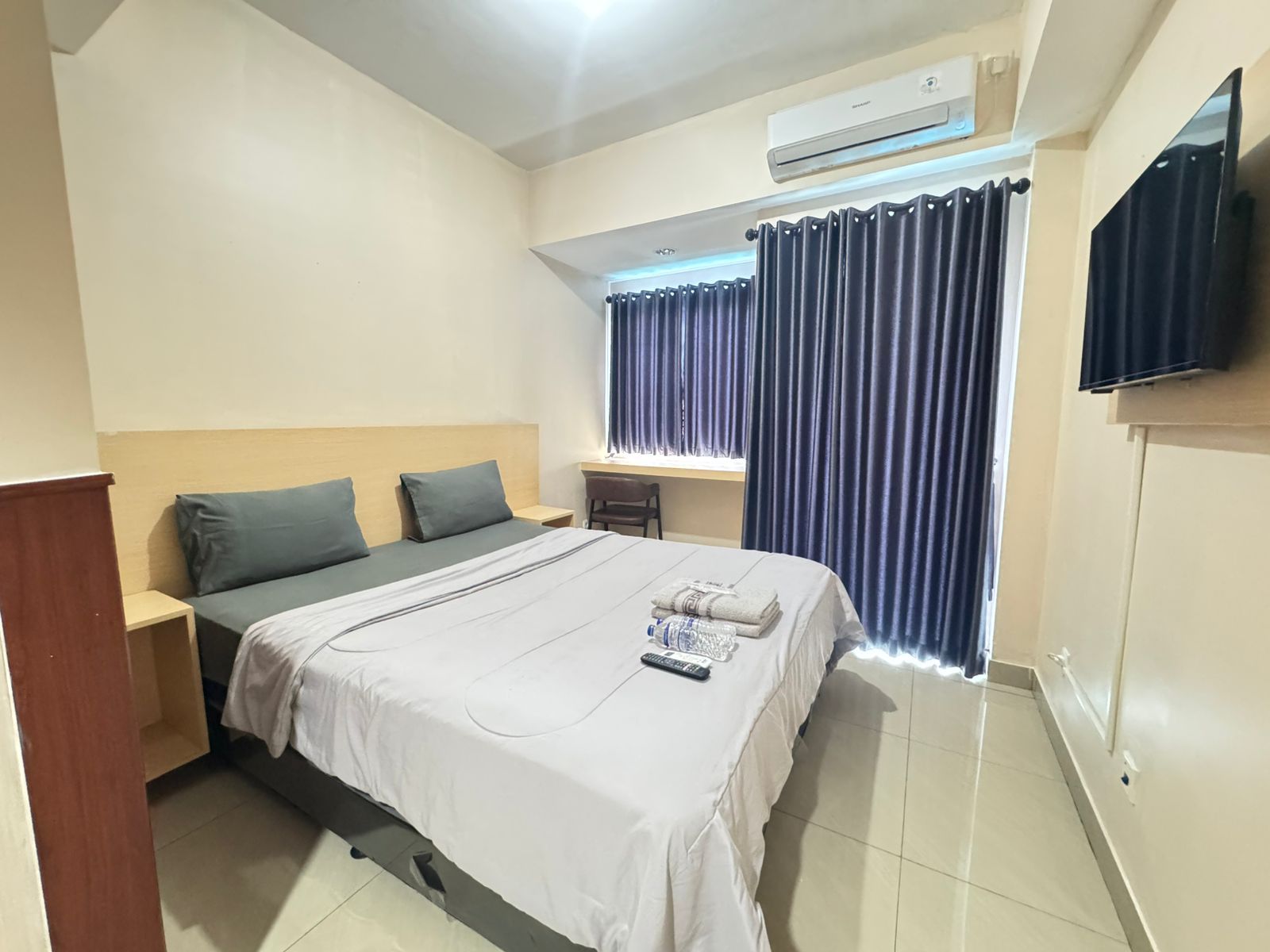 A hotel room with a bed, a desk, an air conditioning unit, a television, and a window with curtains.
