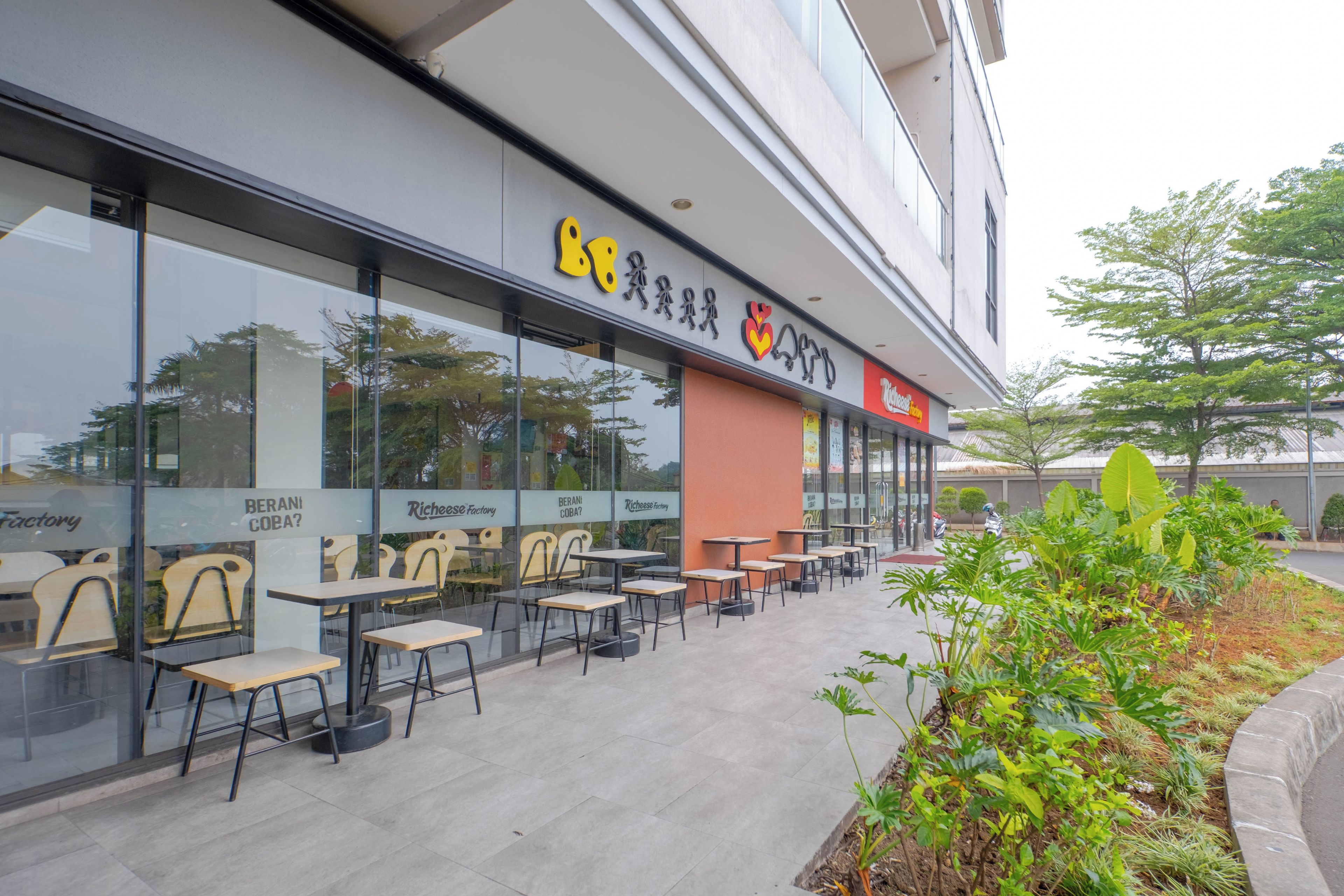 An outdoor seating area with tables and chairs, a glass wall with signage, and a small garden with plants.
