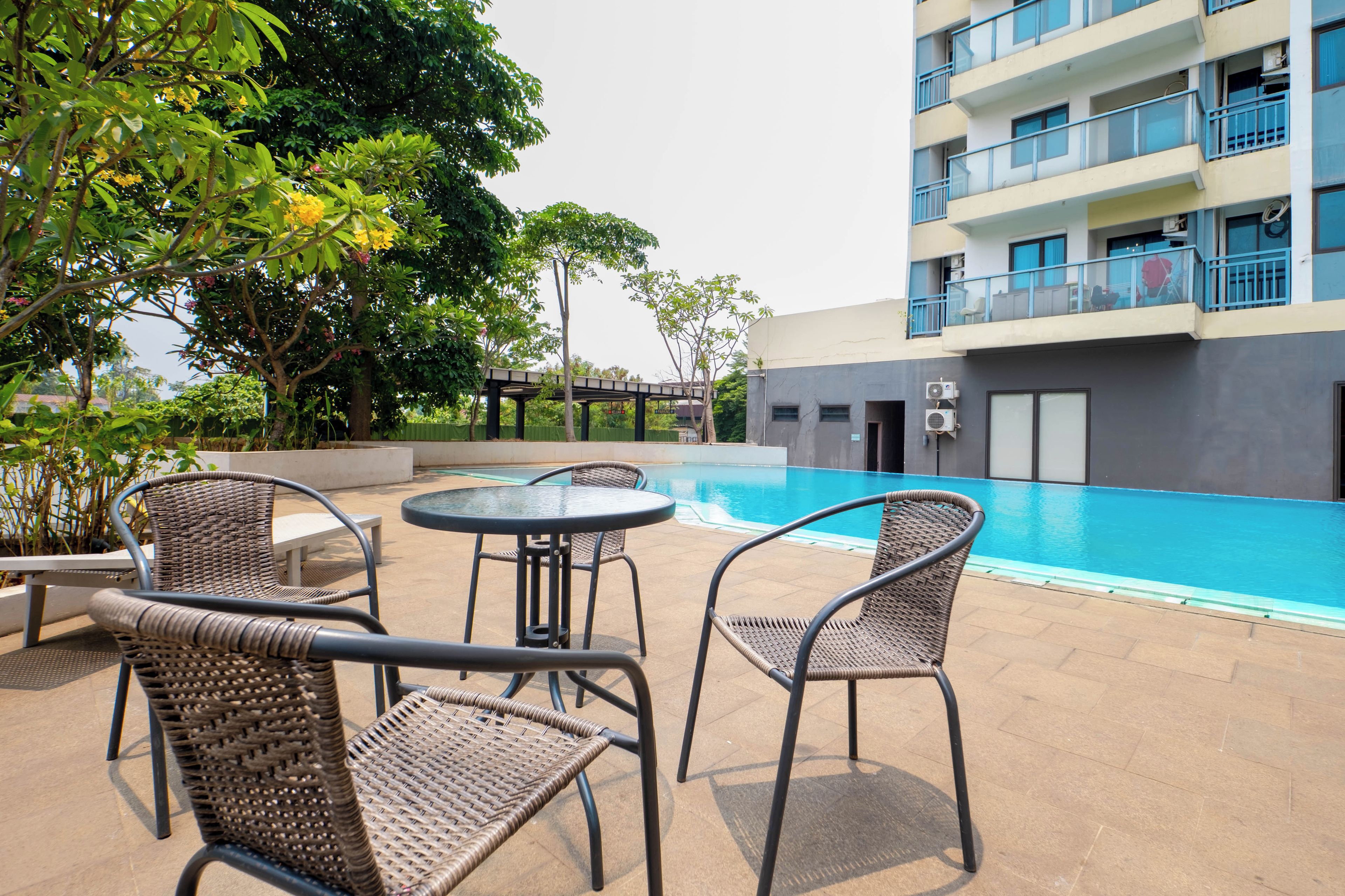 A poolside area with wicker chairs and a table, a building with balconies, and a bridge in the background.