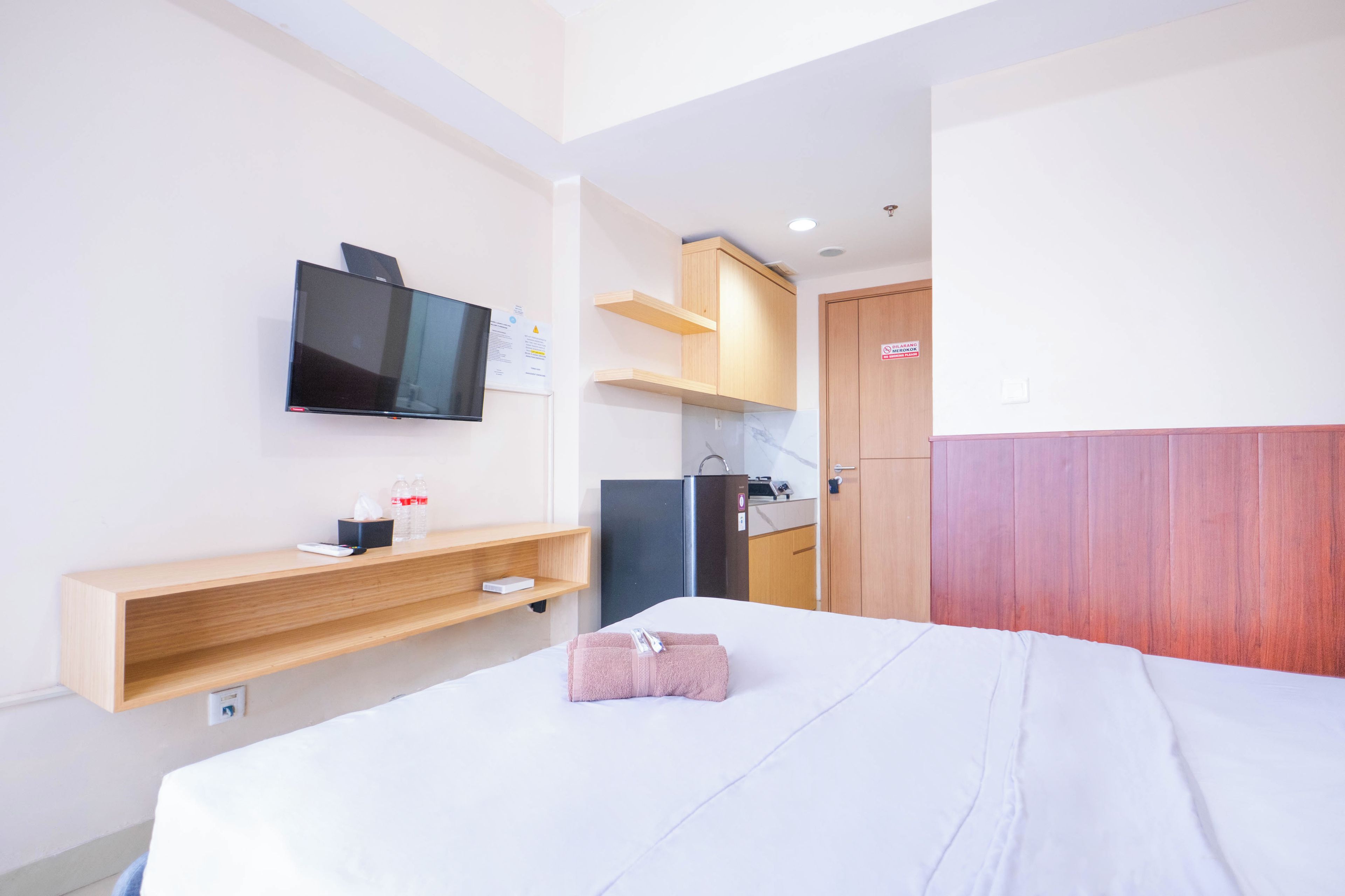 A hotel room with a bed, a television mounted on the wall, a wooden shelf with a remote control, a bottle of water, and a towel.