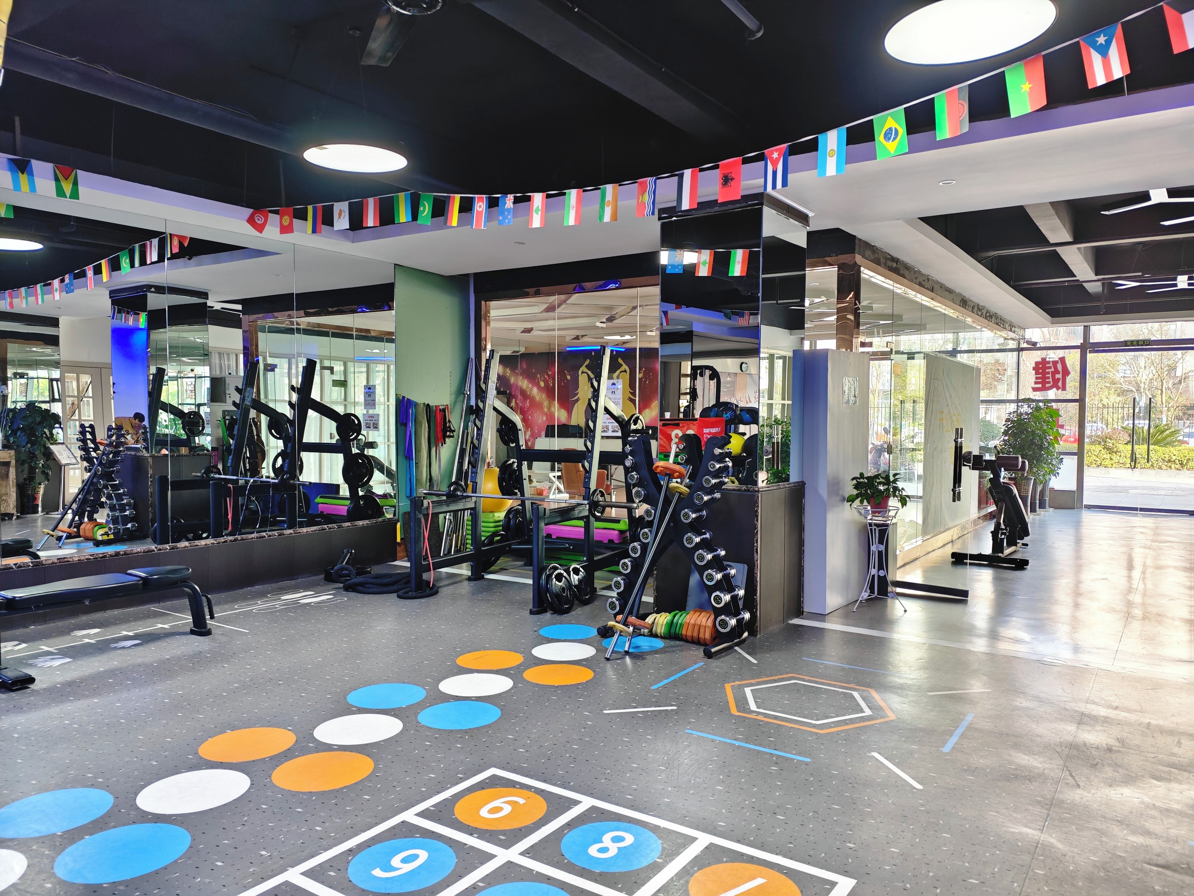 Ein Fitnessraum mit unterschiedlichen Trainingsgeräten, einer Spiegelwand und einem Boden mit nummerierten Kacheln.