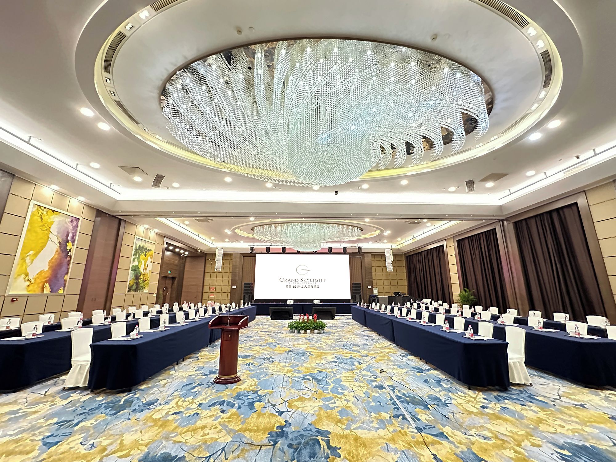 Un salón de conferencias con una pantalla grande, filas de mesas y un techo decorativo.