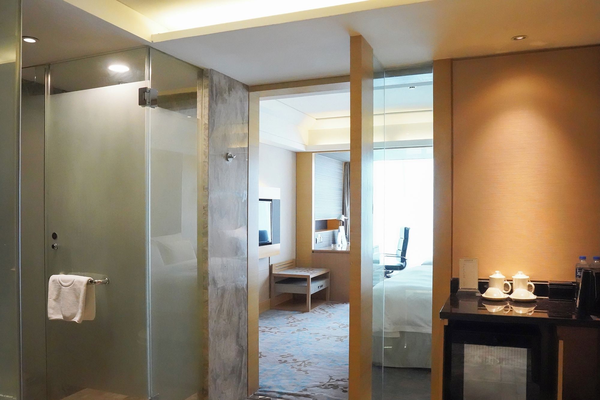 Habitación de hotel con cabina de ducha de vidrio, cama con cabecera, mesita de noche con lámpara, y un pequeño rincón de descanso con una silla y una mesa.