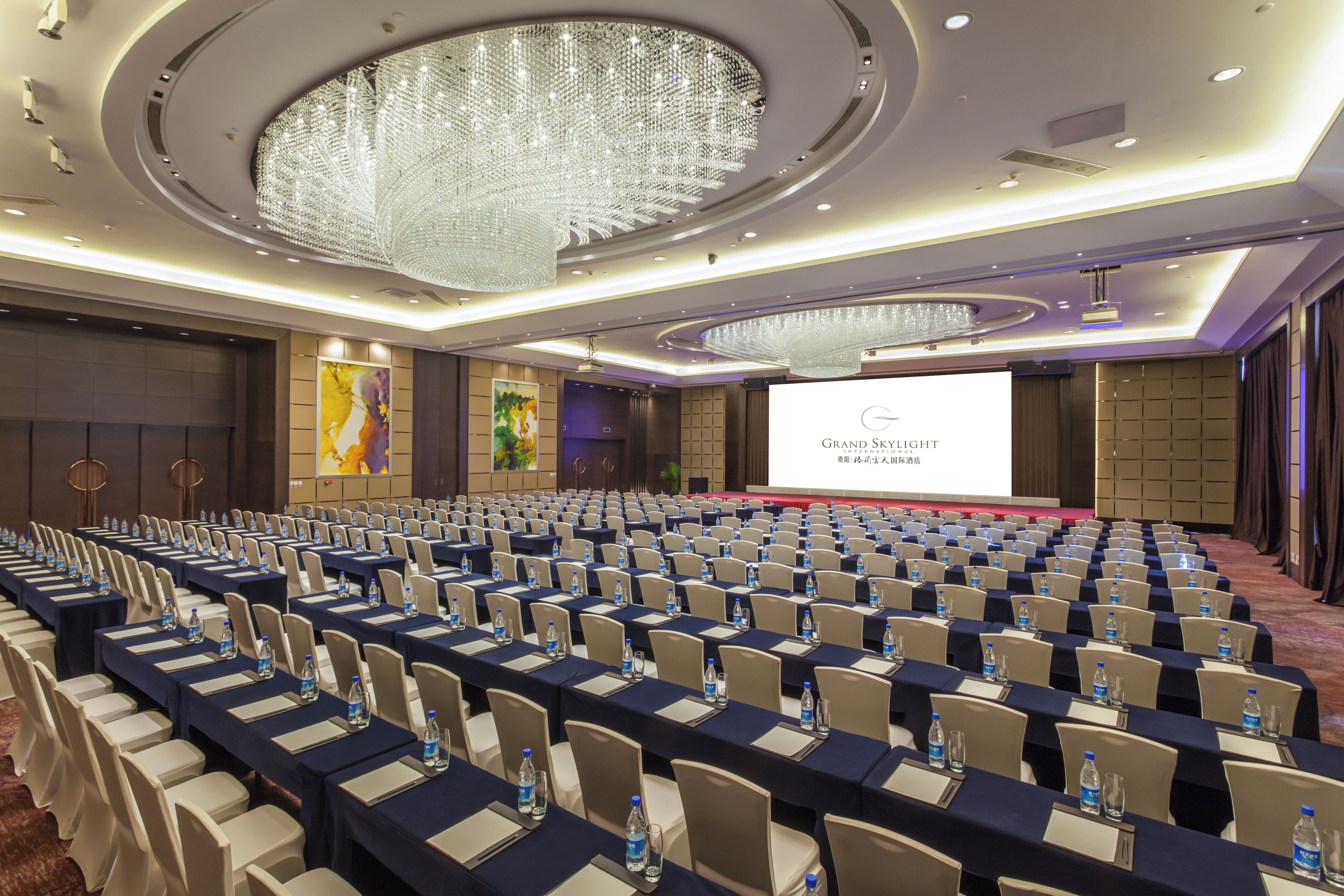 Un salón de conferencias con filas de mesas, sillas y una pantalla grande para presentaciones.