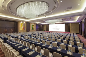 Un salón de conferencias con filas de mesas, sillas y una pantalla grande para presentaciones.