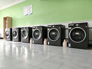 A row of black front-loading washing machines in a commercial setting with a green wall and tiled floor.