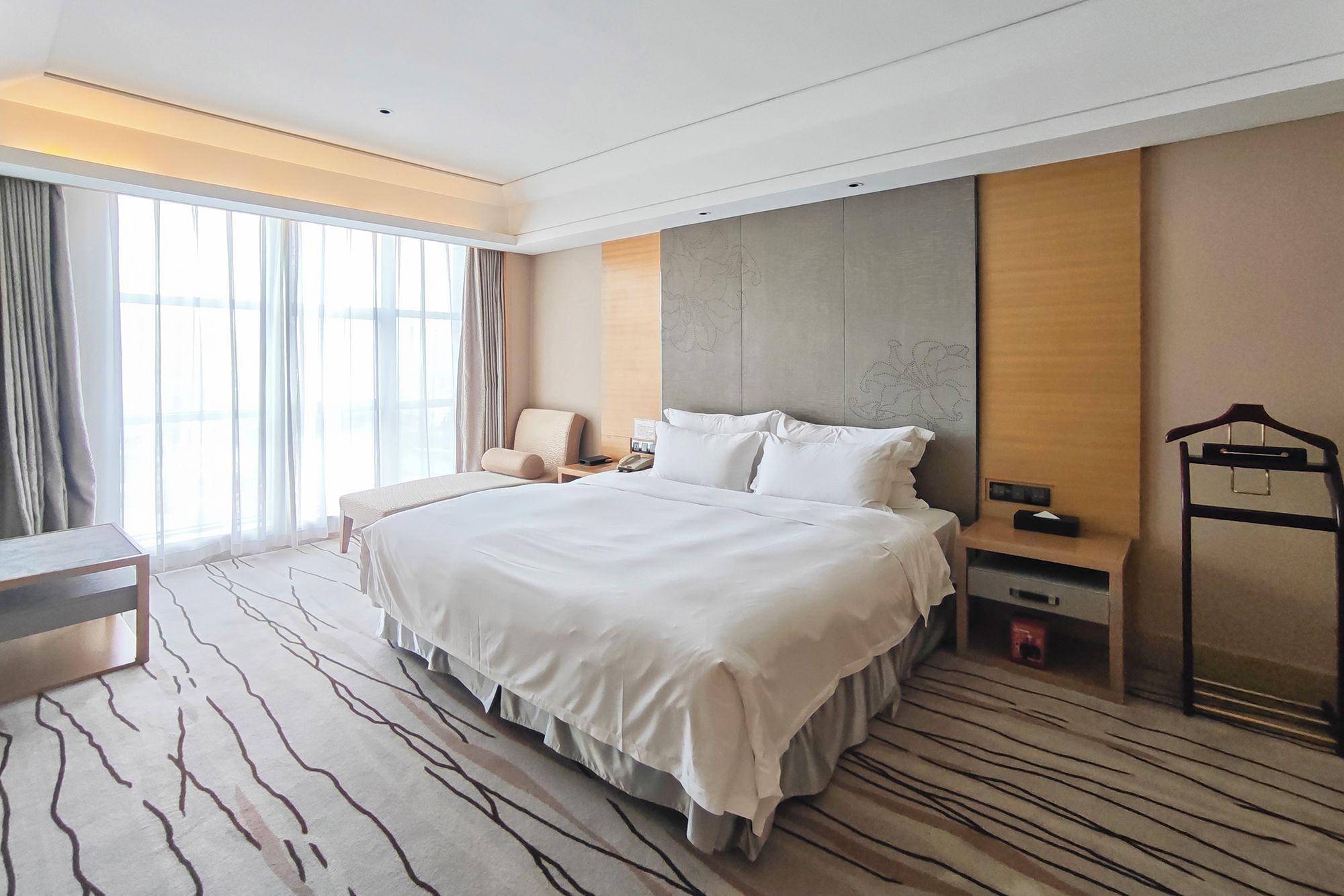 Quarto de hotel com uma cama grande, mesa de cabeceira, cadeira, escrivaninha, guarda-roupa e uma janela ampla com cortinas.