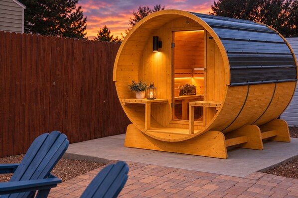 A wooden sauna with a curved roof, a small table with a plant, and two chairs outside.