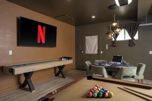 A room with a pool table, pool balls, a foosball table, a television with a Netflix logo, and a poker table.