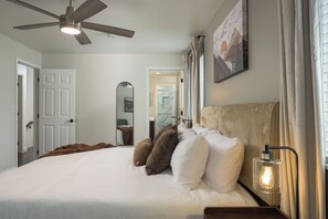 A bedroom with a bed, pillows, a ceiling fan, a nightstand with a lamp, and a painting on the wall.