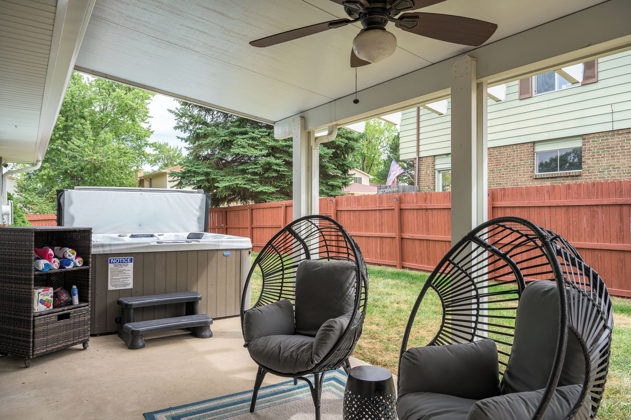 Un espace extérieur abrité, doté d'un ventilateur de plafond, d'un barbecue, de deux fauteuils en osier noirs et d'un espace de rangement sur lequel est apposée une pancarte indiquant « AVIS ».