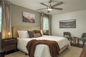 A bedroom with a bed, a ceiling fan, two bedside tables with lamps, a chair, and artwork on the walls.