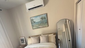 A bedroom with a bed, a nightstand, a wall-mounted air conditioner, a painting, and a full-length mirror. - The Northridge Nook: A Cozy 1-bedroom house in Los Angeles with AC & Pool Access (Los Angeles)