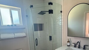 A bathroom with a glass-enclosed shower, a toilet, a sink with a mirror, and a window above the sink. - The Northridge Nook: A Cozy 1-bedroom house in Los Angeles with AC & Pool Access (Los Angeles)