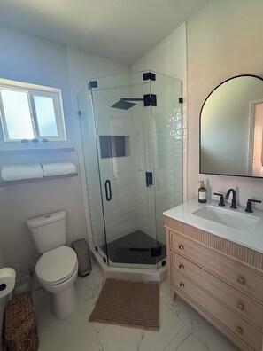 Ein Badezimmer mit einer Duschkabine, einer Toilette, einem Waschbecken mit Spiegel und einem Fenster über dem Waschbecken.