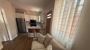 A modern kitchen with white cabinets, a stainless steel refrigerator, and a microwave. There is a dining area with two bar stools and a small table. - The Northridge Nook: A Cozy 1-bedroom house in Los Angeles with AC & Pool Access (Los Angeles)