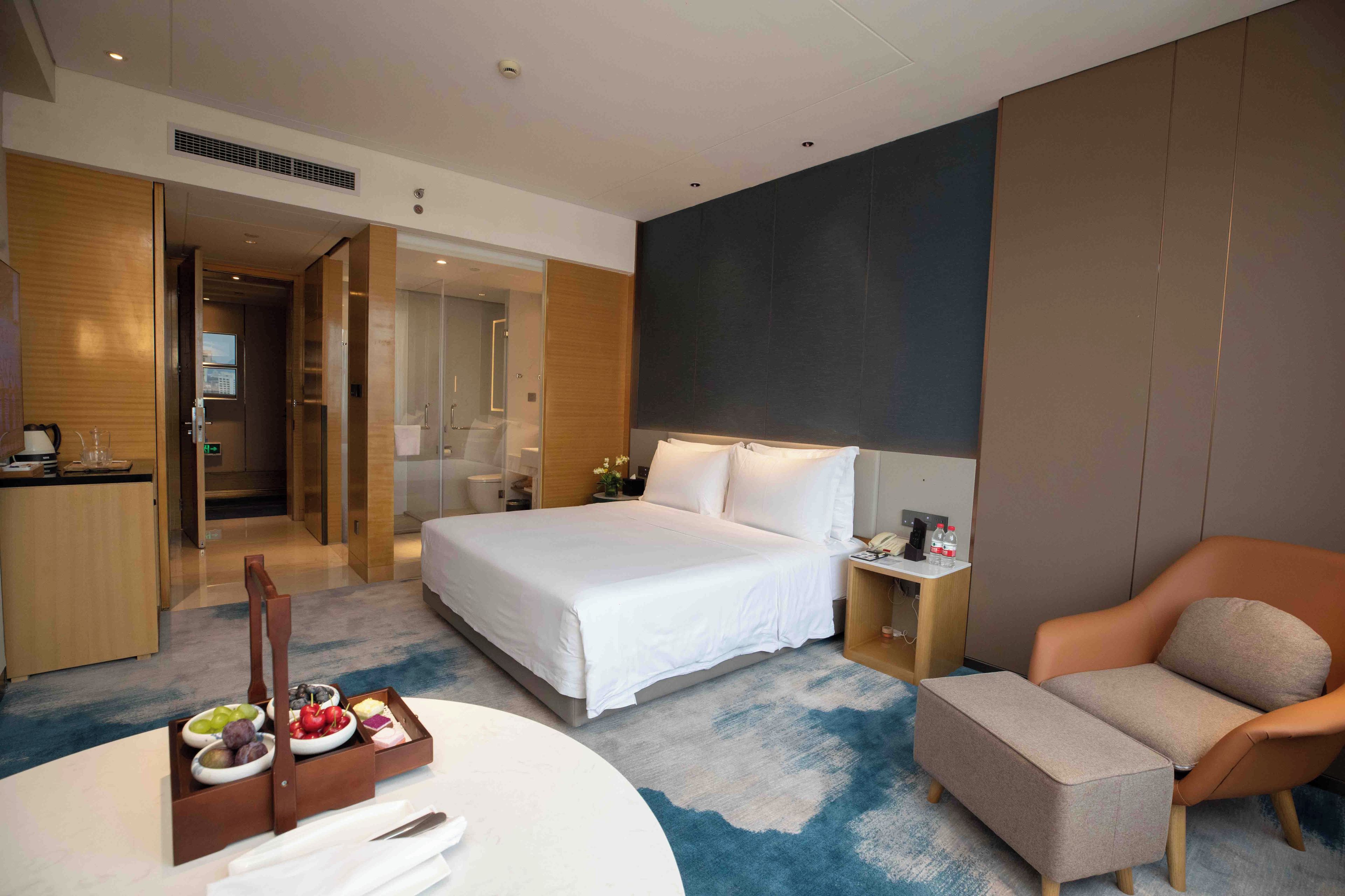 A hotel room with a bed, a desk, a chair, a small table with a fruit tray, and a bathroom visible through an open door.