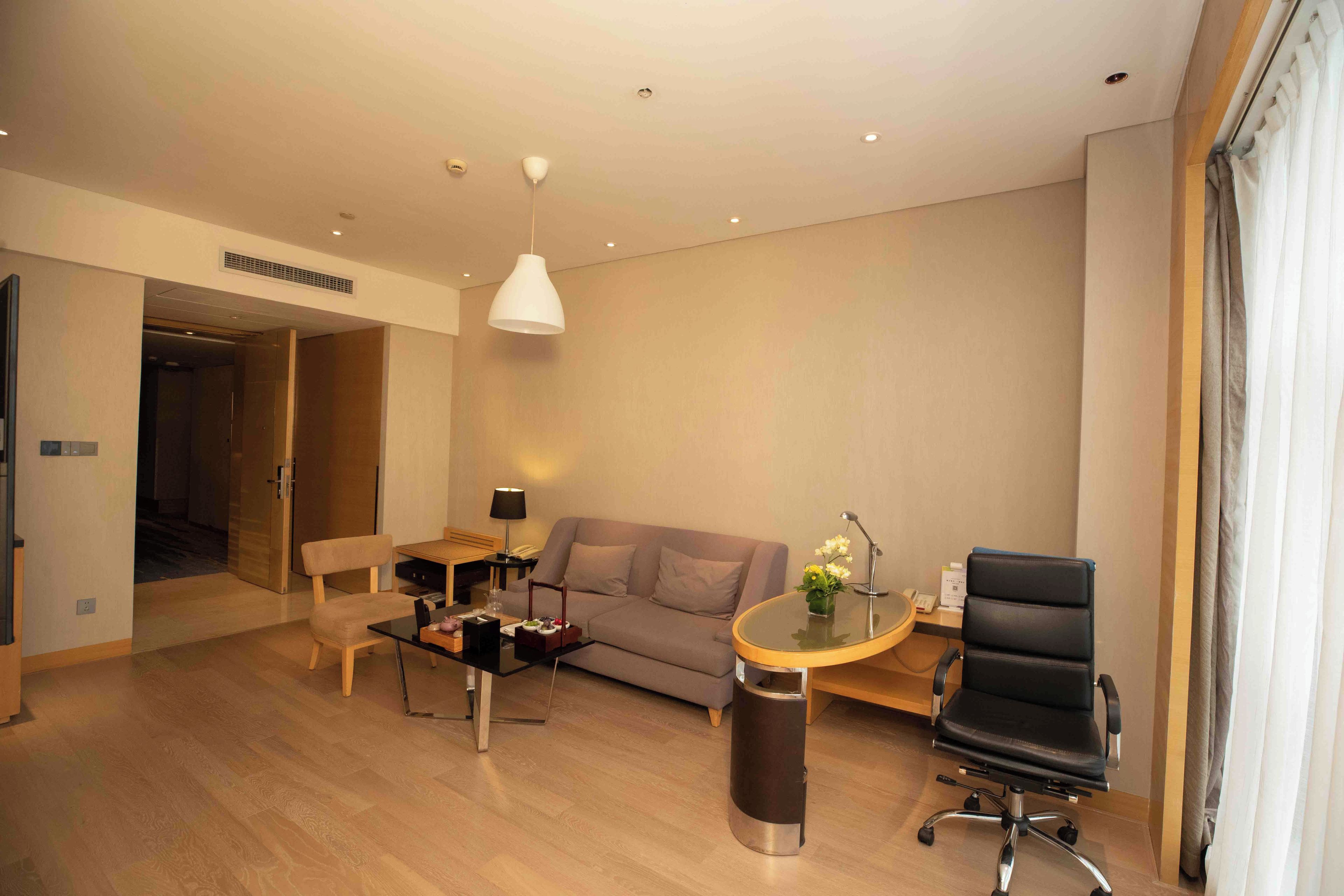 A modern hotel room with a sofa, a dining table, a chair, and a small desk with a lamp.