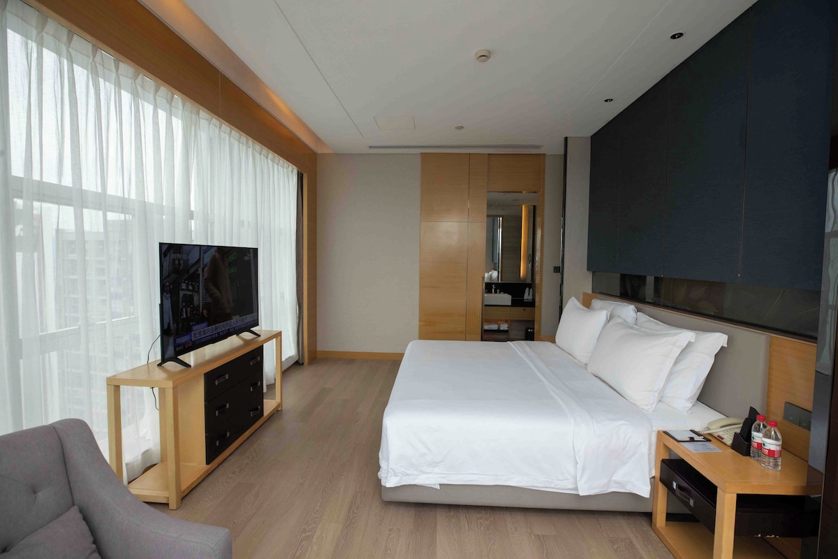 Habitación de hotel moderna con una cama grande, un televisor sobre un soporte de madera, un escritorio con una silla y un ventanal con cortinas.