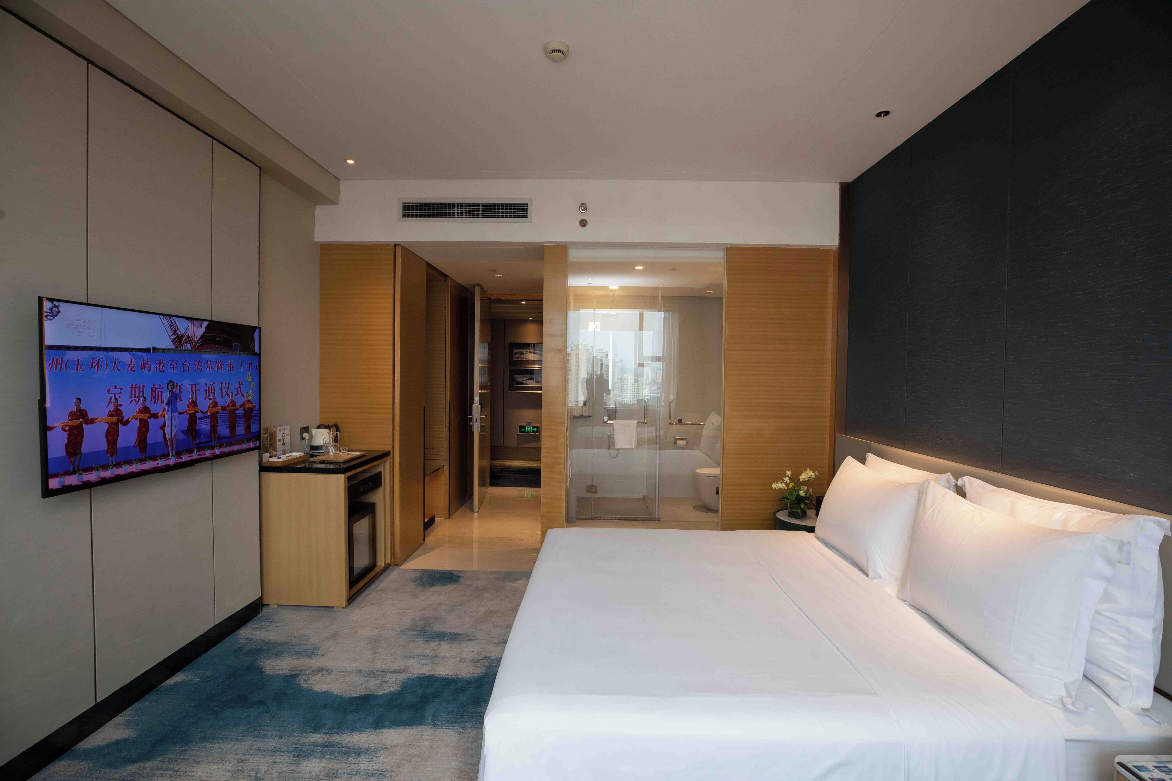 Quarto de hotel com uma cama grande, televisão na parede, uma escrivaninha e um banheiro visível por uma porta aberta.