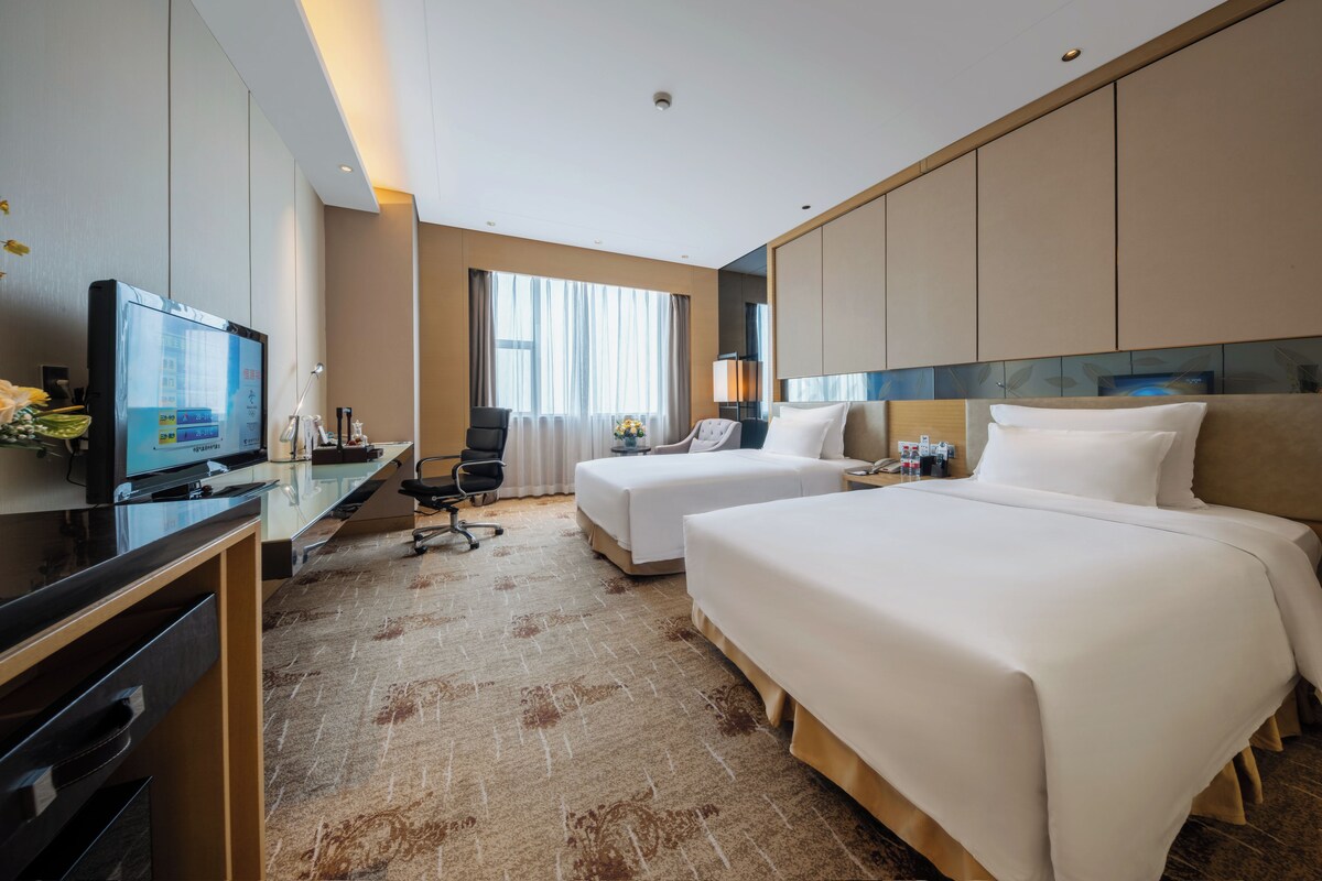 Habitación de hotel con dos camas, un escritorio, un televisor y un ventanal grande.