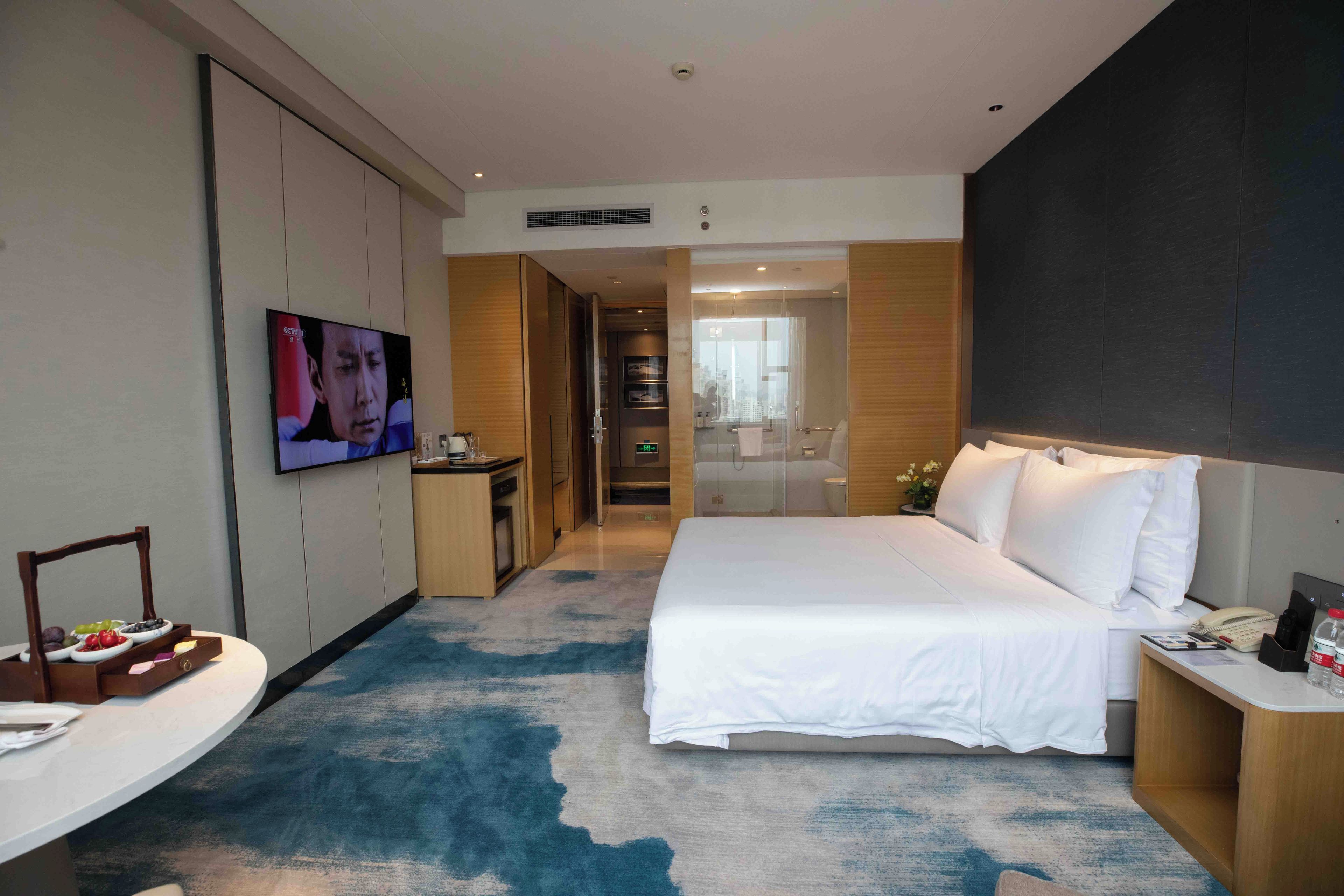A hotel room with a large bed, a TV, a desk with a chair, and a bathroom visible through a glass door.
