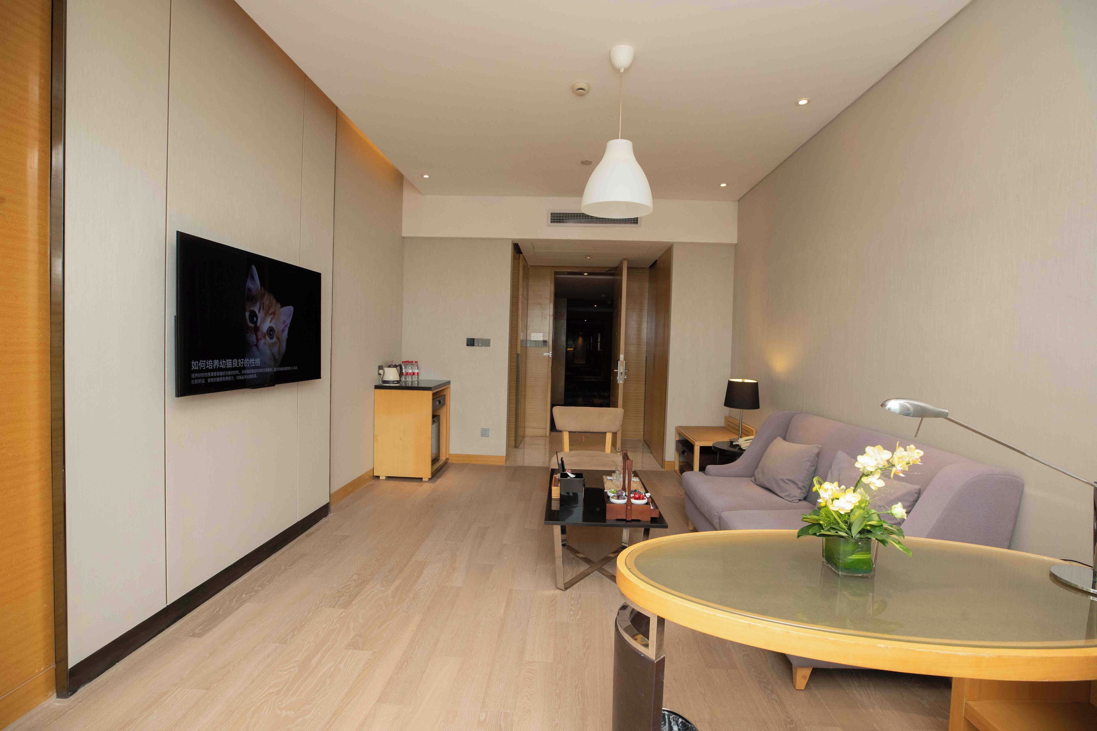 A modern living room with a flat-screen TV mounted on the wall, a round glass coffee table, a sofa, a chair, and a small side table with a lamp.