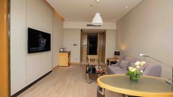 A modern living room with a flat-screen TV mounted on the wall, a round glass coffee table, a sofa, a chair, and a small side table with a lamp.